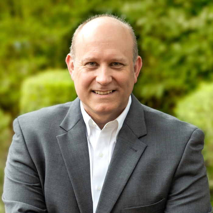 A headshot of John Martinez with green foliage in the background.