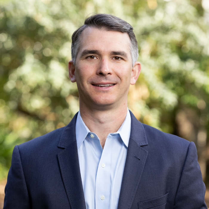 A headshot of John Woody with green foliage in the background.