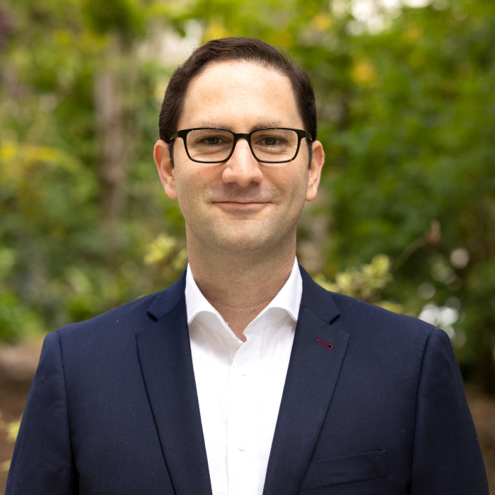 A headshot of Max Gardner with green foliage in the background.